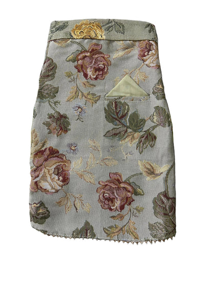 A close-up of a fabric pocket or patch with a vintage floral tapestry pattern in muted pink, green, and beige tones, laid flat on a patterned green and white blanket.