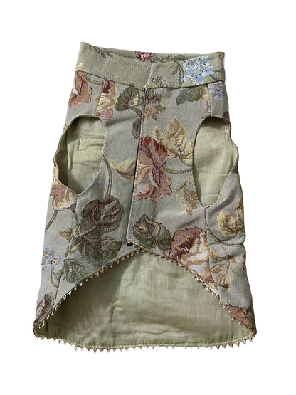 A handmade pet harness made from olive green fabric with a floral pattern in pink, beige, and green, laid flat on a patterned sheet.